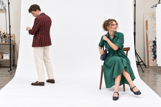 Woman seated in a green midi shirt dress and navy flats while a man checks the cuff of his burgundy striped blazer.