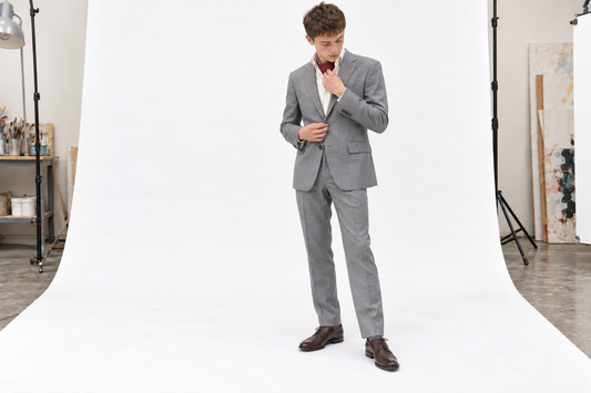 Man adjusting a burgundy day cravat inside a cream shirt collar, with a grey summer suit and brown lace-ups.