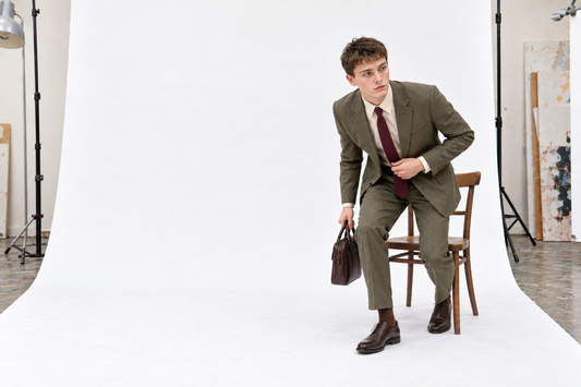 Man rising from a chair in a muted olive summer suit, cream shirt and burgundy tie, carrying a small brown bag.