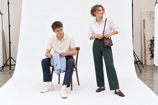 Woman in a white shirt and green trousers beside a seated man in a cream collared knit and navy trousers, preparing practical layers.