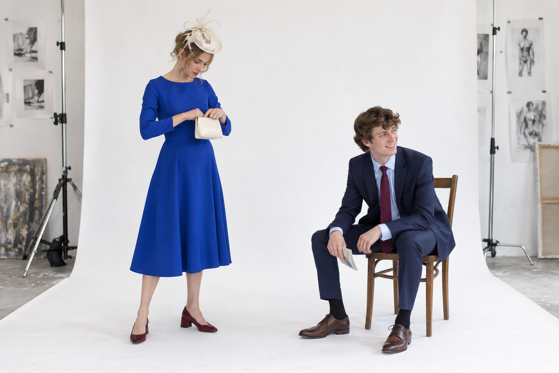 Thackray woman in a cobalt below-knee dress and cream headpiece checking a small clutch while the Thackray man sits nearby in a navy suit with a burgundy tie and folded racecard.