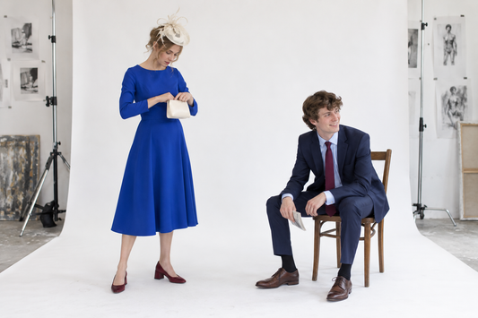 Thackray woman in a cobalt below-knee dress and cream headpiece checking a small clutch while the Thackray man sits nearby in a navy suit with a burgundy tie and folded racecard.