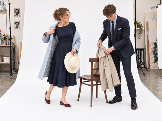 Woman trying a pale-blue coat over a navy dress while a man in a navy suit lifts a stone raincoat from a chair.