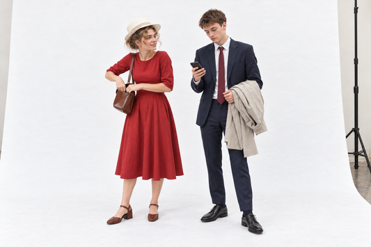 Woman checking a brown shoulder bag in a red sleeved dress and cream hat beside a man checking his phone in a navy suit, carrying a stone raincoat.