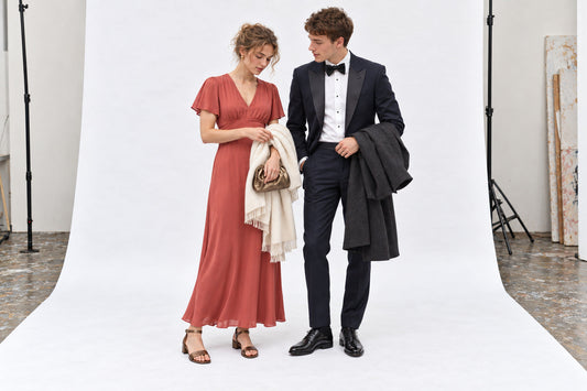 Woman in a coral evening dress and cream wrap beside a man in a midnight-blue dinner suit, in the white studio.