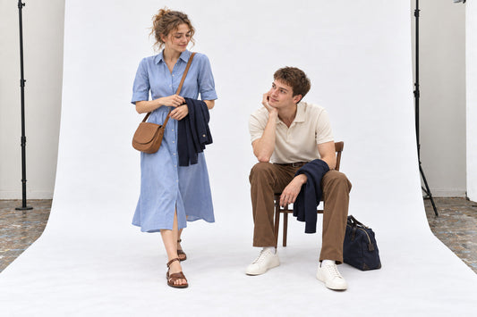 Woman in a blue shirtdress and flat sandals beside a man in an ecru polo and tobacco chinos, in the white studio.