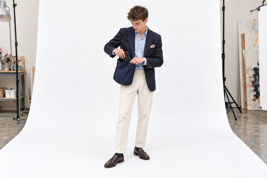 Man packing a phone into a soft navy shoulder bag, wearing a navy jacket, pale blue shirt and stone chinos.