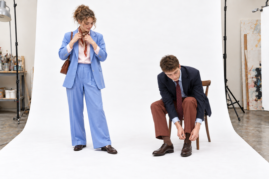Woman adjusting a rust silk scarf with a cornflower suit beside a seated man tying his shoes in a navy blazer and rust trousers.