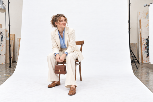 Woman settling into a chair in a cream trouser suit and blue shirt, holding a small structured bag above tan loafers.